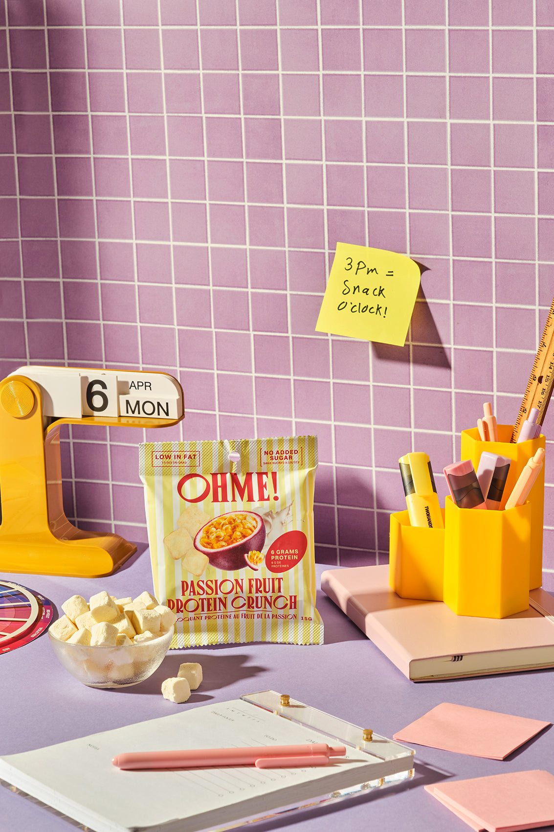 OHME! passion fruit snack pack on a desk with stationery items and a purple tiled wall.
